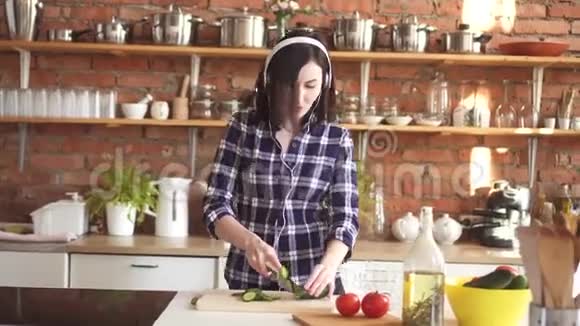 穿着衬衫和耳机的女人在厨房里听音乐跳舞切蔬菜视频的预览图