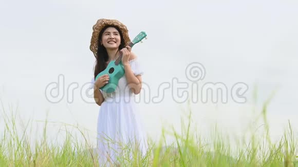 亚洲快乐的女人穿着白色连衣裙在绿色的草地上弹着四弦琴吉他夏天有着山山的背景音乐美人视频的预览图