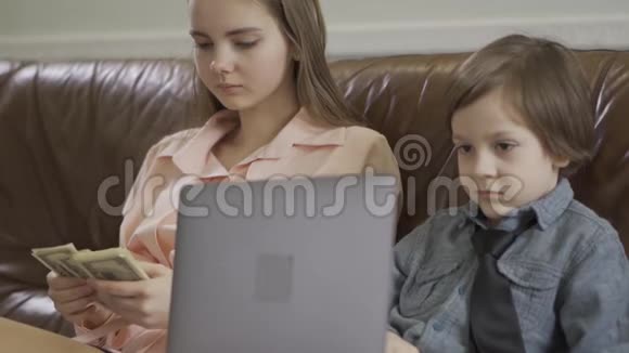 姐姐和弟弟坐在皮革沙发上那个男孩拿着笔记本电脑女孩在数钱视频的预览图