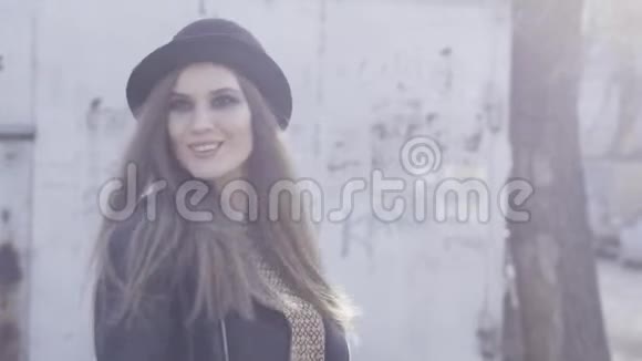漂亮的时髦女孩带着黑色时尚的帽子和皮夹克在温暖的阳光下摆在老建筑里行动微笑视频的预览图