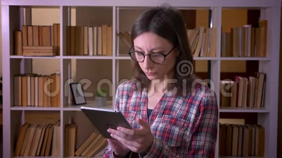用平板电脑和图书馆里的照相机拍摄一位年轻漂亮的女学生戴眼镜的特写镜头视频的预览图