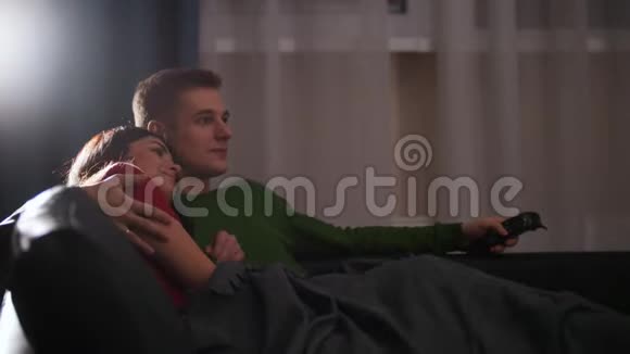 年轻可爱的一对夫妇躺在沙发上拥抱在一起看电视视频的预览图