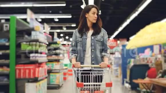 年轻的头发蓬乱的黑发女郎正在超市里散步推着购物车去买食物四处张望视频的预览图