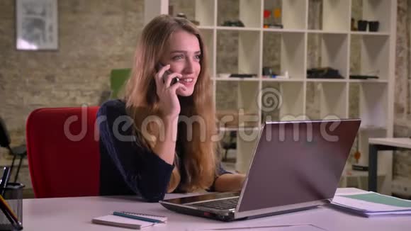 白种人金发女商人在办公室笔记本电脑前愉快地用手机交谈的肖像视频的预览图