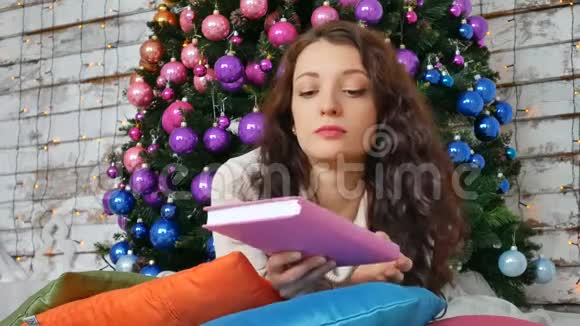 美丽的女人背景是圣诞树在明亮的枕头上看书女孩子的画像视频的预览图