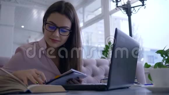 在线教育戴眼镜的学生女孩在现代上网本上的视频聊天中在笔记本上写笔记视频的预览图