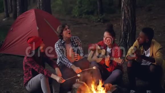 年轻的女旅行者正在弹吉他她的朋友们正在着火做饭非裔美国人正在鼓掌视频的预览图