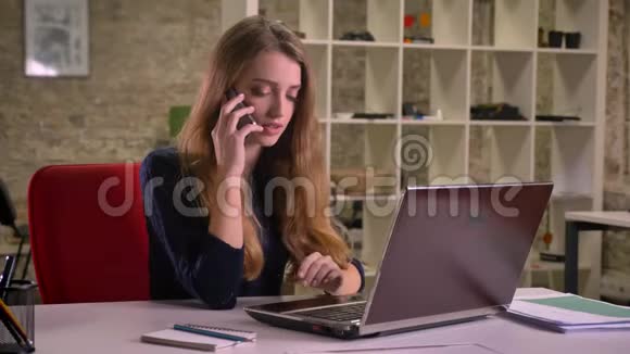 白种人金发女商人在办公室的笔记本电脑前认真地用手机说话的肖像视频的预览图