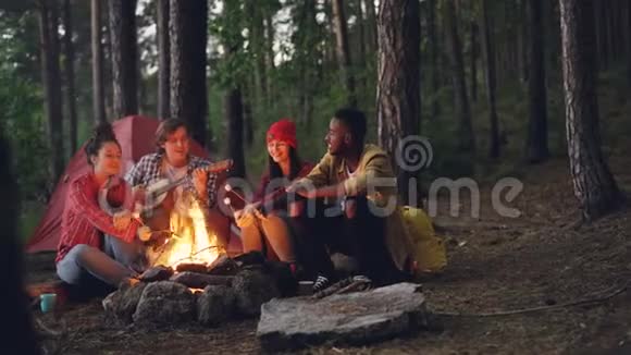 轻松的男女游客们在森林里的篝火旁唱歌弹吉他烤棉花糖视频的预览图