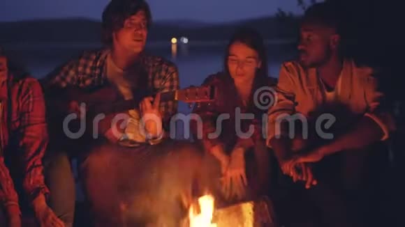 快乐的朋友们在夏天的夜晚在河边的森林里唱着歌弹着吉他取暖视频的预览图
