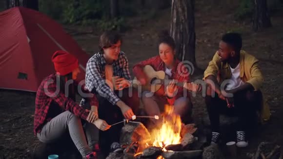 漂亮的女人正在弹吉他而她的朋友们正在唱着滑稽的歌笑着在火上煮棉花糖视频的预览图