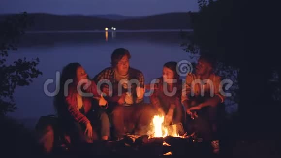 在湖边森林的篝火旁快乐而放松的男孩和女孩在唱歌而他们的朋友在弹吉他视频的预览图