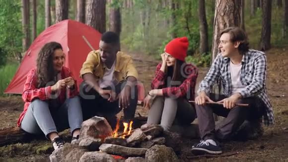 快乐的游客男人和女人分享坐在篝火旁的故事笑着在燃烧的火焰附近取暖视频的预览图