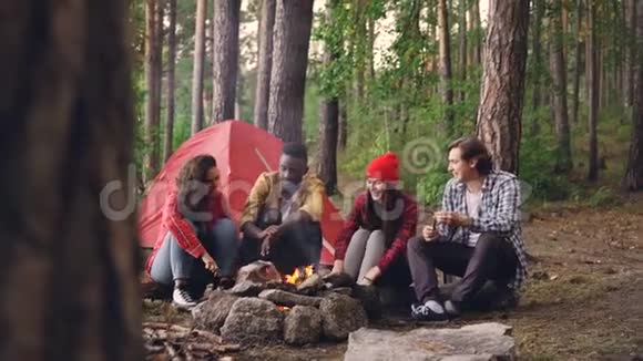 快乐的女孩和男人朋友们在营地里聊天打手势笑着围着火炉休息取暖视频的预览图