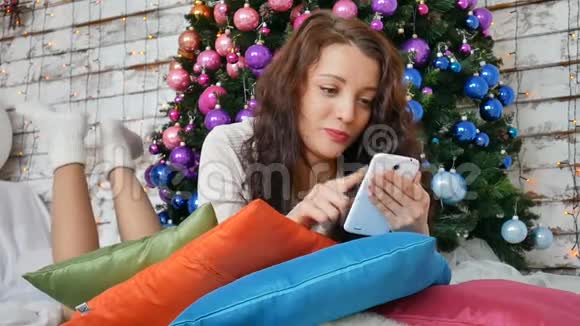 卷曲的黑发女人躺在一棵圣诞树旁的枕头里圣诞树上装饰着粉红色的紫色球她还和自己一起上网视频的预览图
