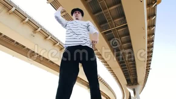一位年轻的波普戴着帽子穿着一件条纹T恤在夏天在桥下跳舞展示自由泳视频的预览图