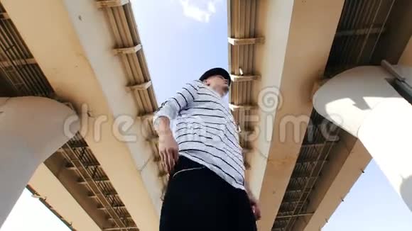 在炎热的夏日一个戴着帽子穿着条纹T恤的年轻人在桥下翩翩起舞视频的预览图