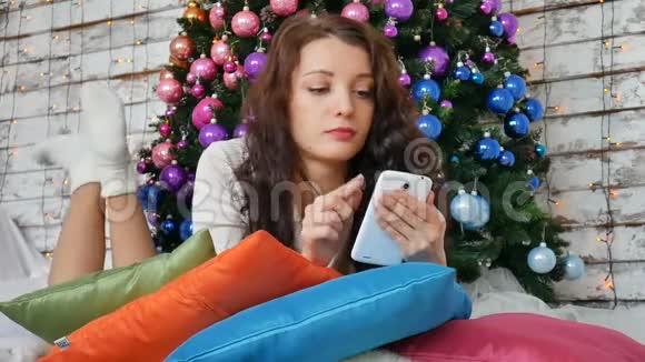 女孩躺在地板上手里拿着电话在家里的圣诞树背景上视频的预览图