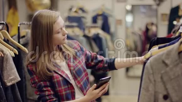 成年美女在服装店的货架上寻找一件新衣服手里拿着智能手机视频的预览图
