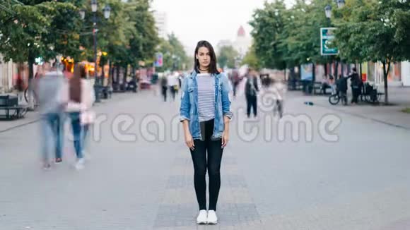 紧张的年轻女子独自站在市中心穿着牛仔裤和牛仔夹克看着她的照片视频的预览图