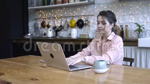 年轻女子专心致志地坐在厨房的木桌旁用她的银色笔记本电脑和一杯视频的预览图