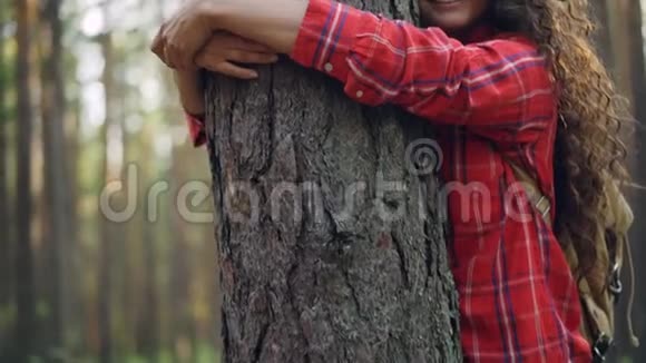 美丽的年轻女子一头卷发穿着鲜艳的衬衫拥抱着树享受着大自然闭着眼睛微笑着视频的预览图