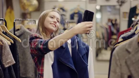 快乐的年轻女性购物者试着穿上一件衣服穿着一件衣服在智能手机摄像头上拍照视频的预览图