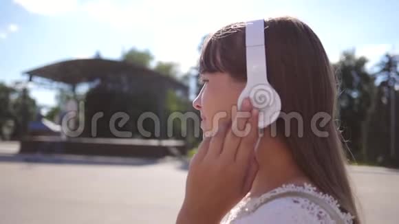 女孩带着耳机沿着城市街道行走听音乐和微笑穿着白色长裙子的女孩视频的预览图