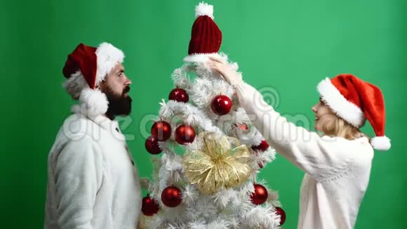 幸福的夫妇在绿色的背景上装饰一棵圣诞树戴着新年帽子的大胡子男人和金发女人玩得很开心视频的预览图