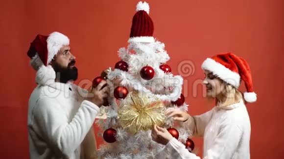 年轻夫妇在红色背景上装饰圣诞树戴着顶帽子的大胡子男人和美女视频的预览图