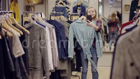 一位年轻漂亮的幸福女人在一家服装店里用她的智能手机相机拍摄自己的照片视频的预览图