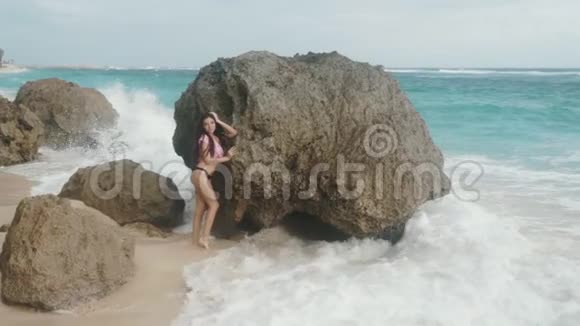 慢动作漂亮的年轻女孩穿着泳衣站在大海和海浪背景下的大石头附近稳重的视频的预览图