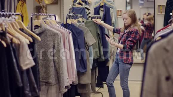 一位迷人的年轻女士拿着智能手机在服装店的货架上检查衣服视频的预览图