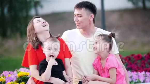 幸福的家庭在公园的花朵中小儿子坐在妈妈腿上女儿坐在爸爸腿上视频的预览图