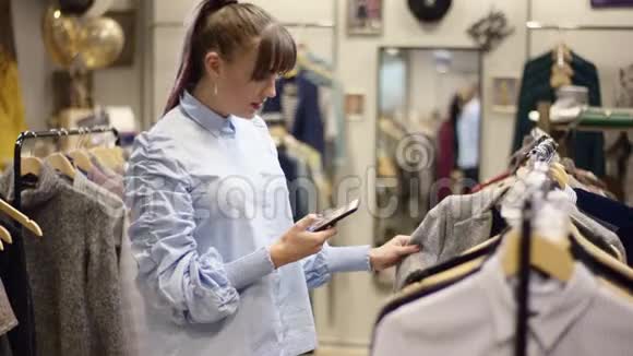 一个漂亮的成年女人在服装店里用智能手机相机拍照一边挑选衣服视频的预览图
