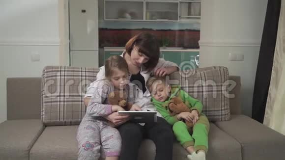 快乐的妈妈和两个孩子坐在沙发上女人向孩子们展示平板电脑家庭假日视频的预览图