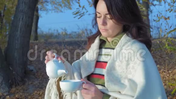 清晨坐在长凳上的一位坐着瓷茶壶绿茶的美丽女子视频的预览图