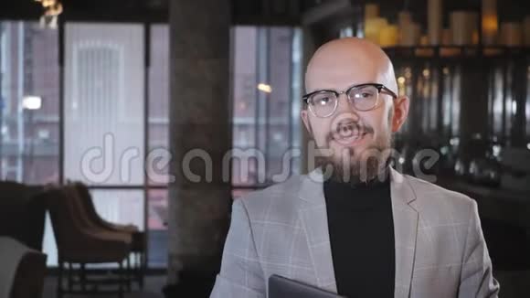 有胡子的年轻人的肖像戴着一套眼镜看着摄像机在咖啡馆里对着镜头微笑视频的预览图