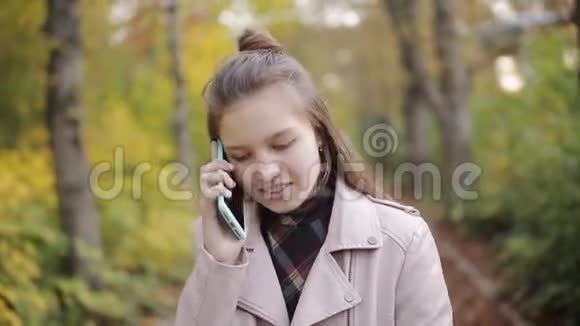 迷人的女孩少年n带着一本书在手机上聊天在美丽的秋天公园散步快乐女孩视频的预览图