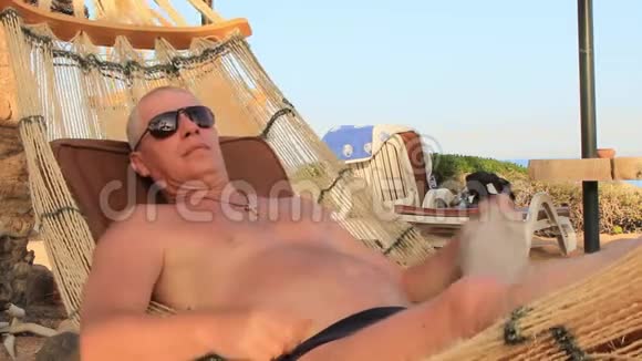 一个阳光明媚的夏日戴着太阳镜的年轻人在海滩上的吊床上休息视频的预览图