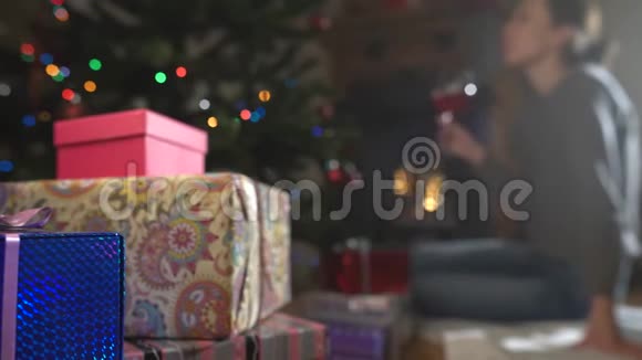 年轻女子正在装饰圣诞树附近和壁炉旁喝酒重点放在礼物上视频的预览图