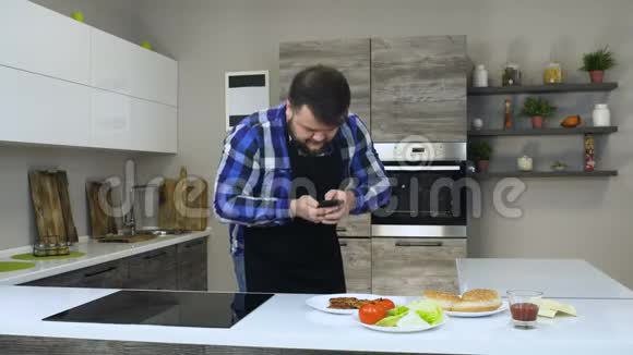 一个长着大胡子的胖子在做一个自制的汉堡之前在厨房里用智能手机视频的预览图