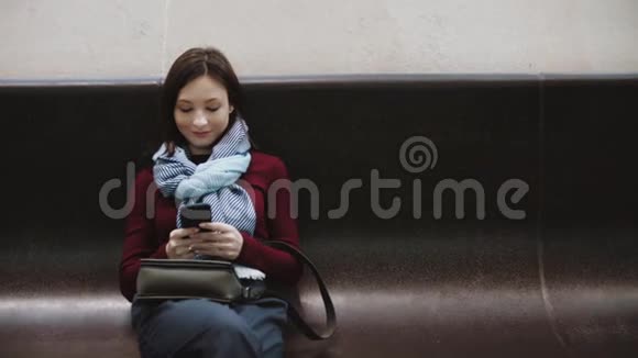 兴奋的女孩在坐在长凳上的智能手机上发短信在车站等火车视频的预览图
