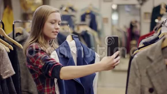 成年迷人的微笑女人带着菲托在智能手机摄像头上穿着一件衣服从一个架子上拿出来的夹克视频的预览图