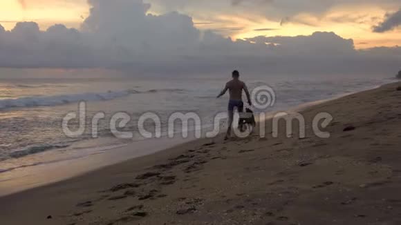 日落时分一个年轻人牵着一条狗在热带海洋的海滩上奔跑视频的预览图