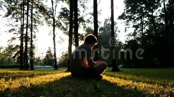 一个人用电话在公园的草地上坐着日落时举起双手视频的预览图