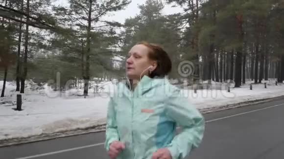 年长的白种人女人戴着耳机在冬天的雪地公园里跑步关闭前面跟随射击慢动作视频的预览图