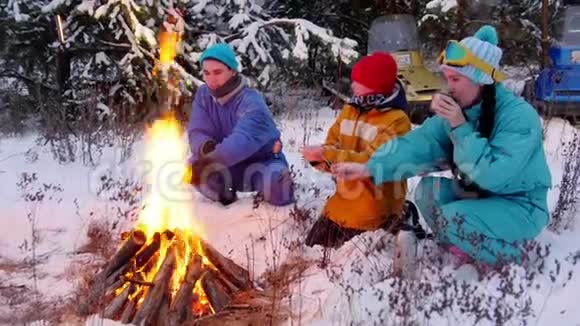 冬季森林一家人坐在火边的树林里一个年轻的女人喝热水瓶里的茶视频的预览图