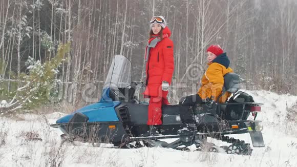 冬天一个穿着五颜六色夹克的年轻女子和小男孩站在雪地摩托车旁视频的预览图