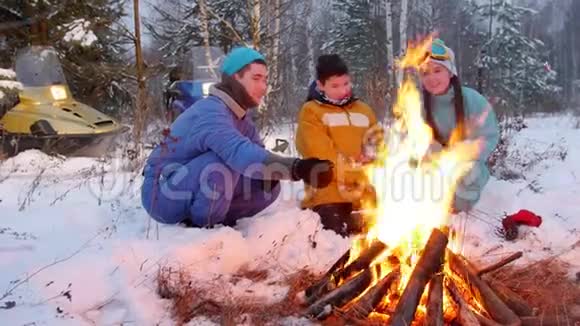 冬季森林一家人坐在火边的树林里把香肠烤成肉串视频的预览图